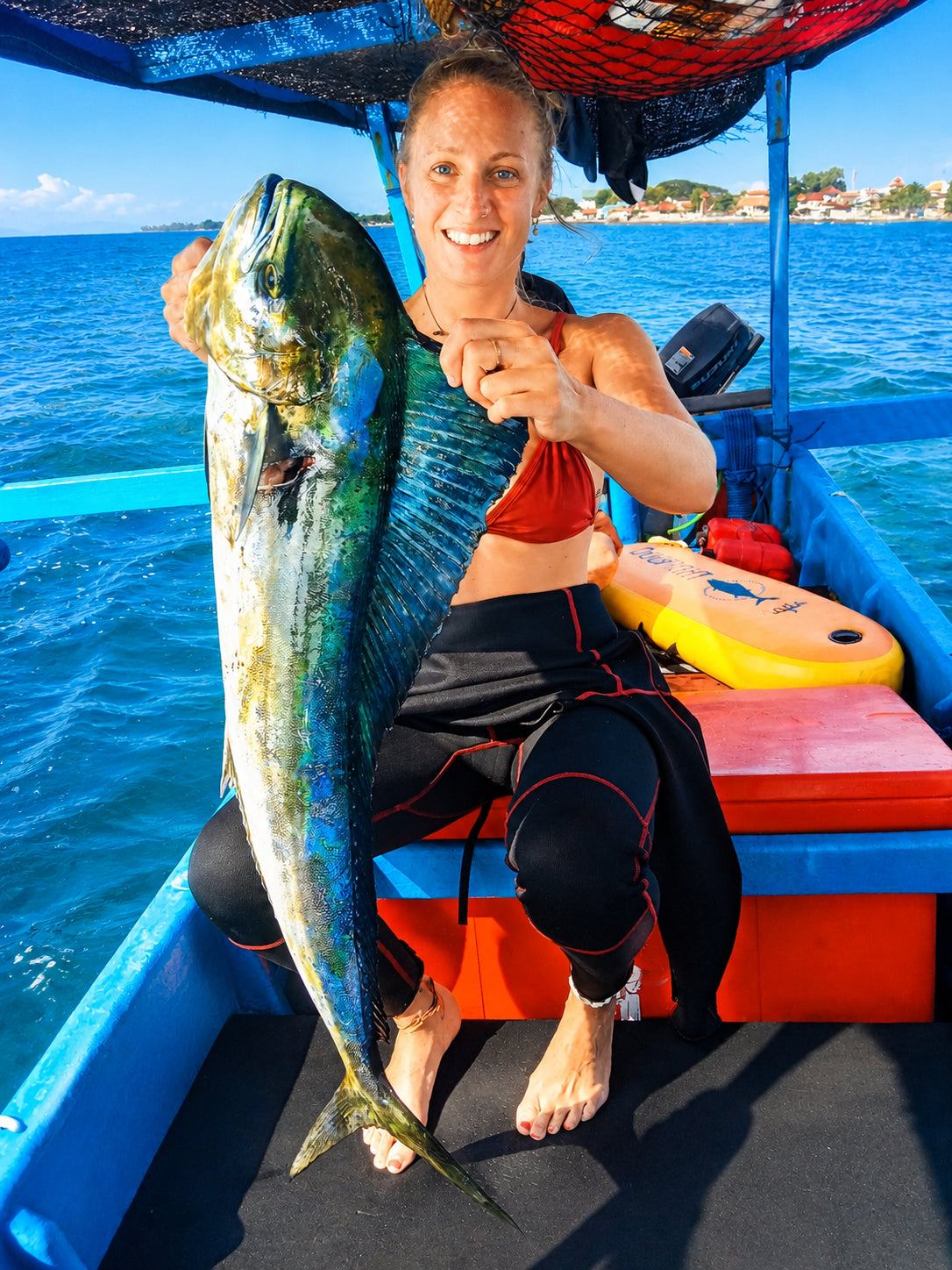 Girl in a wetsuit sitting on an ice box hold up a large size mahi mahi fish that was recently speared in the North Bali Sea