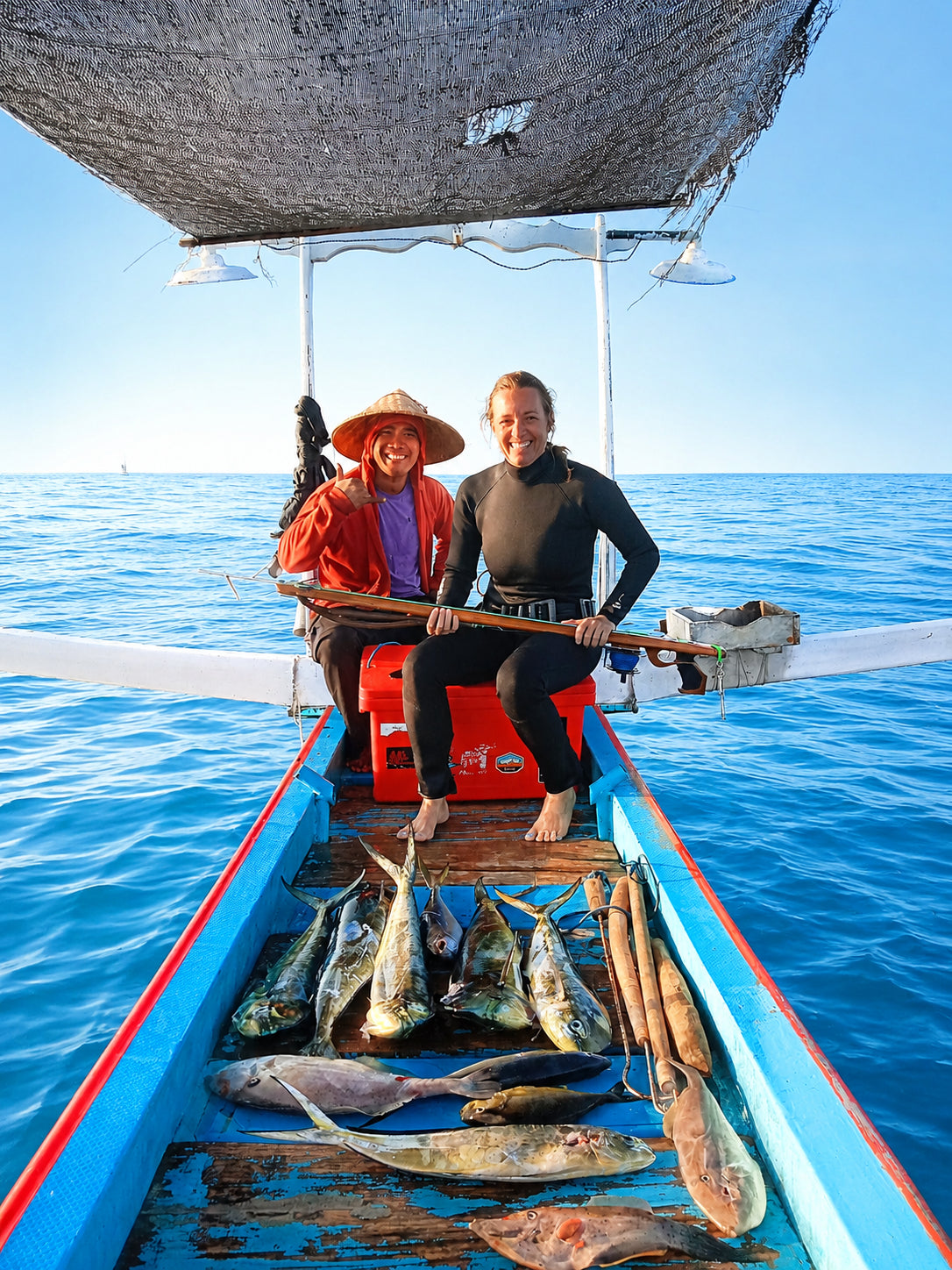 Woman in a wetsuit holding a speargun next to a local boat driver displaying 8 fish on the deck of the boat that were speared in Bali 