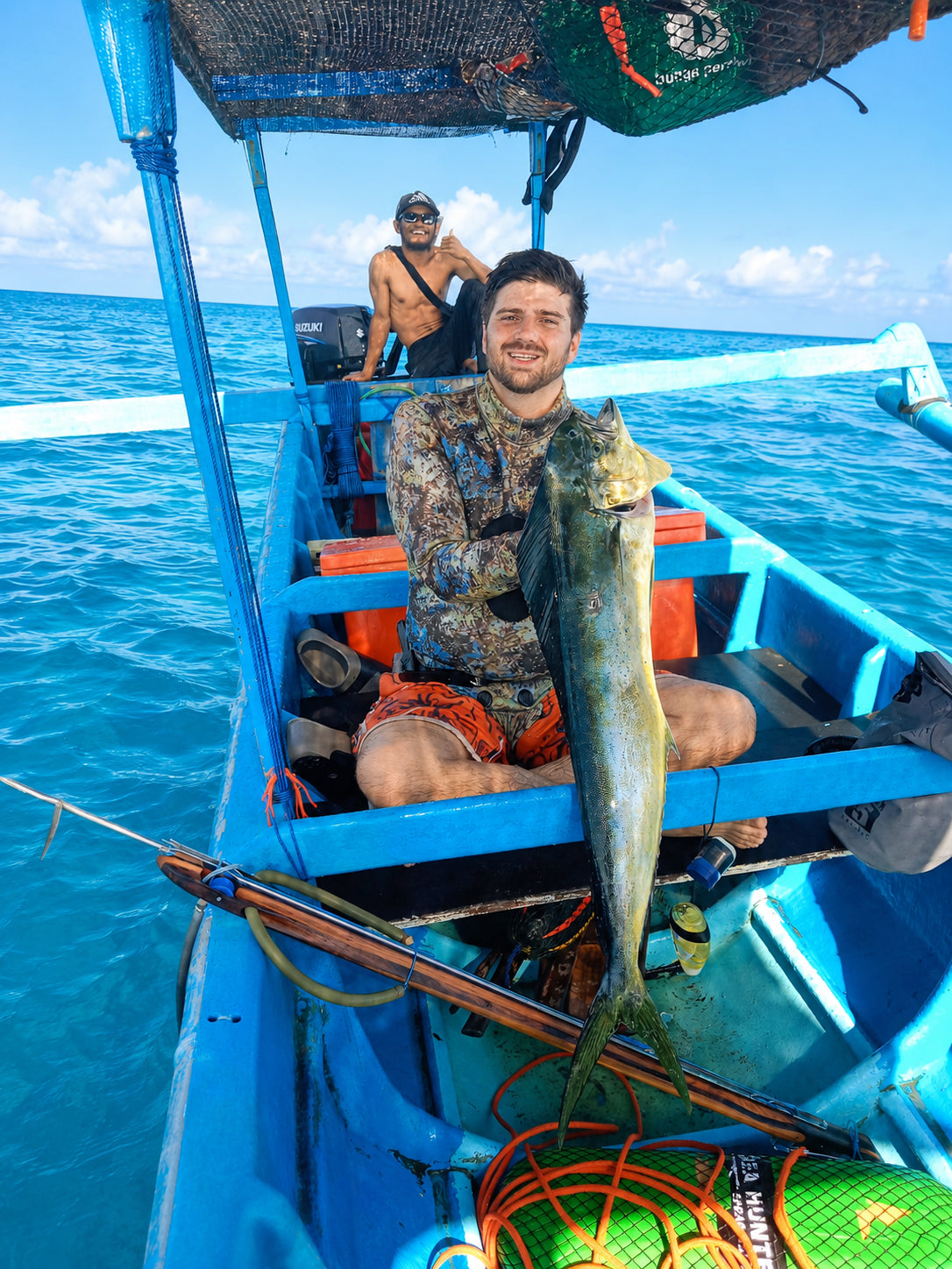 Man holding up a large mahi mahi fish while sitting on a blue local Balinese boat offshore with clear weather and flat ocean conditions