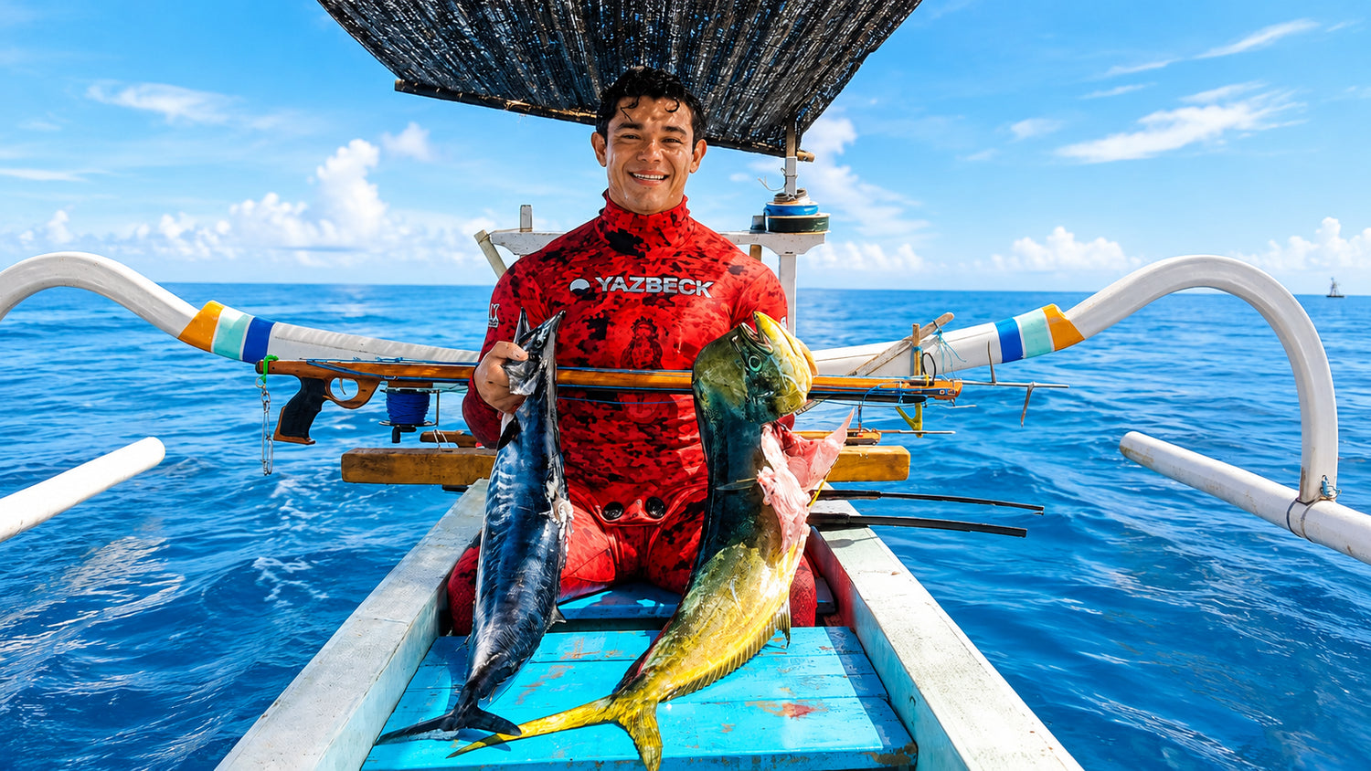 Man in a red wetsuit sitting on a Balinese local boat holding a speargun and a mahi and a wahoo fish that was just speared