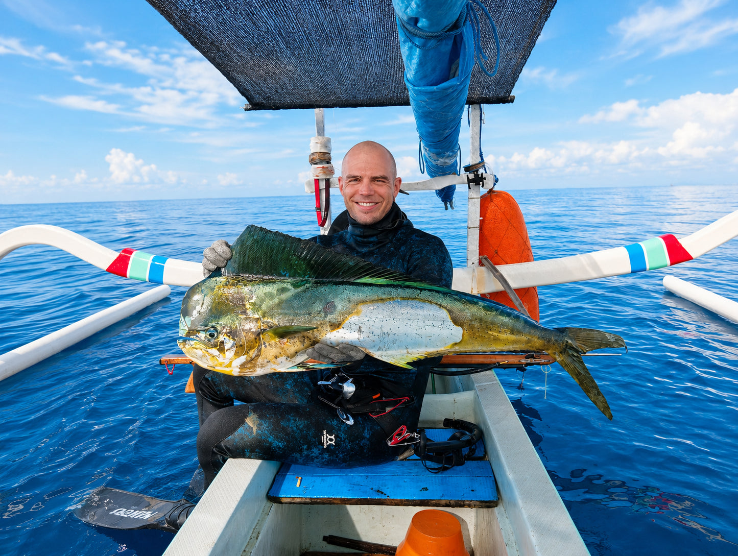 Man sitting on a Balinese jukung boat holding a large mahi mahi fish he just spearfished offshore in Amed Bali